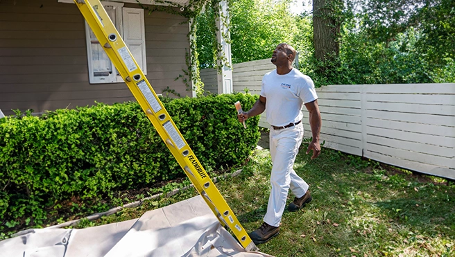 Painting professional holding paintbrush beside ladder outside of residential home.